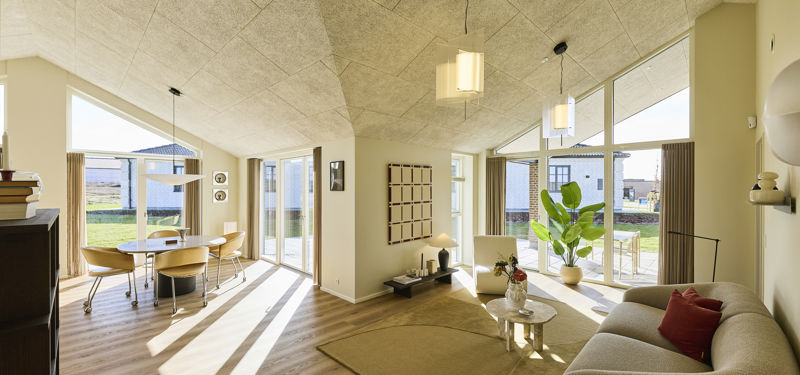 Åbent opholdsrum fra eurodan-huse med stue og spiseplads i samme rum, store vinduespartier, skrå akustiklofter, lys sofa, rundt sofabord, grøn plante og spisebord ved terrassedøren – inspiration til nybyggeri og husdrømme med sammenhængende køkken-alrum.