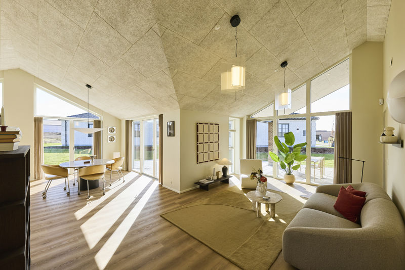 Åbent opholdsrum fra eurodan-huse med stue og spiseplads i samme rum, store vinduespartier, skrå akustiklofter, lys sofa, rundt sofabord, grøn plante og spisebord ved terrassedøren – inspiration til nybyggeri og husdrømme med sammenhængende køkken-alrum.