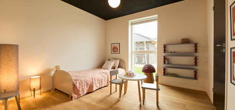 Børneværelse fra eurodan-huse med enkeltseng med lyserødt sengetæppe, lille rundt bord, børnestol, lave væghylder, gulvlampe og højt vindue med dagslys – inspiration til børneværelse i nybyggeri, husbyggeri og moderne husdrømme.