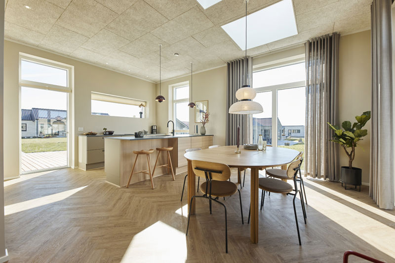 Køkken-alrum fra eurodan-huse med spiseplads ved rundt træbord, pendel over bordet, lang køkkenø med barstole, store gulv-til-loft-vinduer, høje gardiner, ovenlys og sildebensgulv – inspiration til nybyggeri med dagslys, opholdszoner og køkkendrømme.