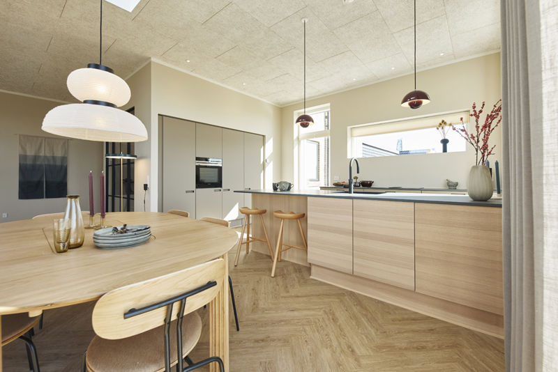 Lyst køkken-alrum i moderne vinkelhus med træfronter, stor køkkenø, spisebord, barstole, pendellamper og ovenlys – inspiration til nybyg og drømmehuset.