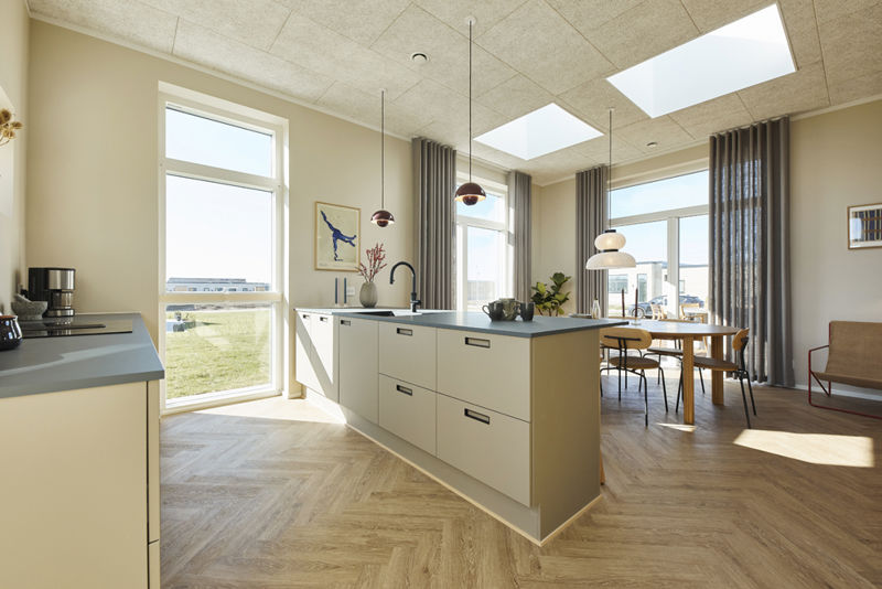 Lyst køkken-alrum fra eurodan-huse med lang køkkenø i lyse fronter, mørk bordplade, sort armatur, barstole i træ, to ovenlysvinduer, store vinduer med gardiner og spiseplads ved rundt bord – inspiration til nybyggeri med naturligt lys og moderne køkkenløsninger.