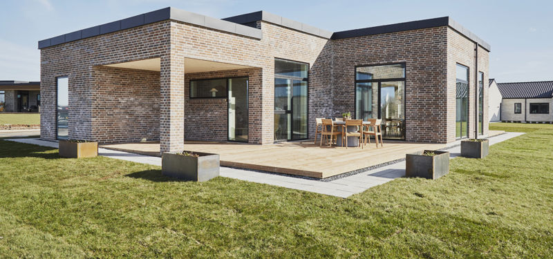 Stor træterrasse fra eurodan-huse placeret mellem lyse murstensfacader med overdækket hjørne, brede glaspartier, udendørs spiseplads og firkantede plantekasser i græsset – inspiration til moderne nybyggeri, terrassemiljø og husdrømme.