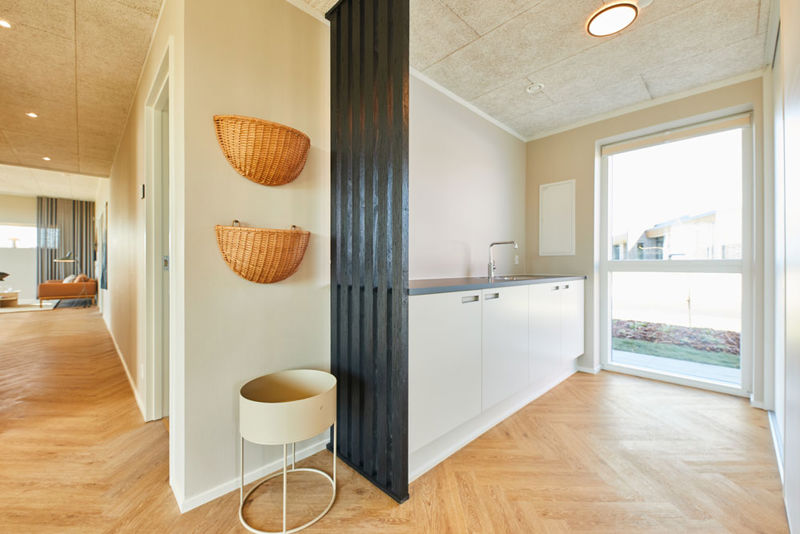 Lyst bryggers i moderne vinkelhus med hvide skabe, lang bordplade, stort vindue, sildebensgulv og sort lamelvæg – inspiration til praktisk indretning af nybyg og drømmehuset.