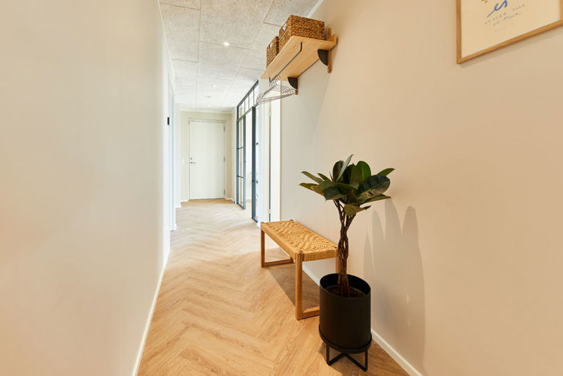 Lys gang i moderne vinkelhus med sildebensgulv, træbænk, væghængt knagerække og grøn plante – inspiration til praktisk indretning af nybyg og drømmehuset.