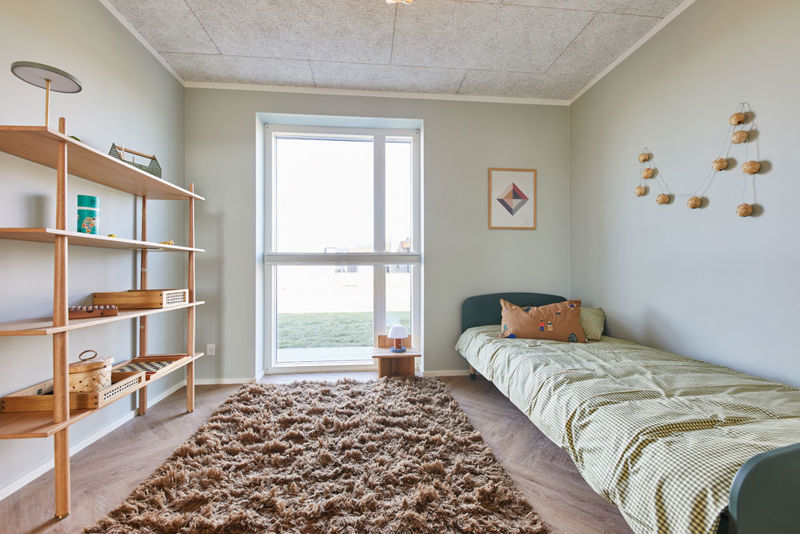 Hyggeligt børneværelse i moderne vinkelhus med enkeltseng, væghængt reol, stort vindue og blødt gulvtæppe – inspiration til indretning af nybyg og drømmehuset.