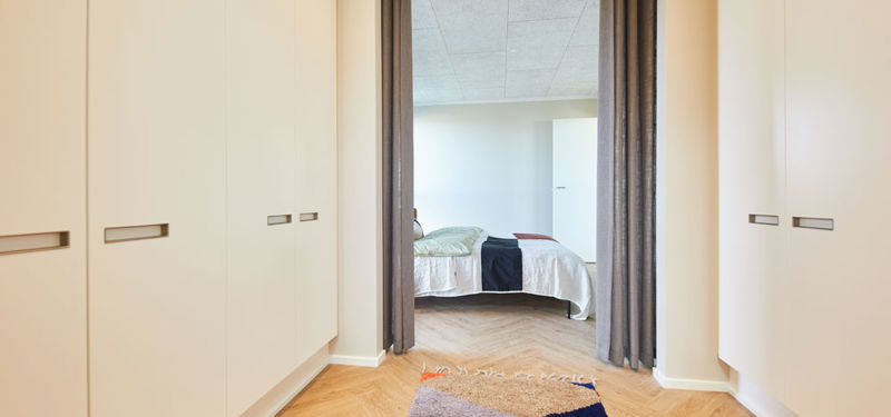Walk-in passage fra eurodan-huse med lyse skabsvægge på begge sider, lange integrerede greb, gardiner som rumdeling, farvet gulvtæppe og kig ind til soveværelse med seng – inspiration til garderobe mellem rum i nybygget hus og moderne husbyggeri.