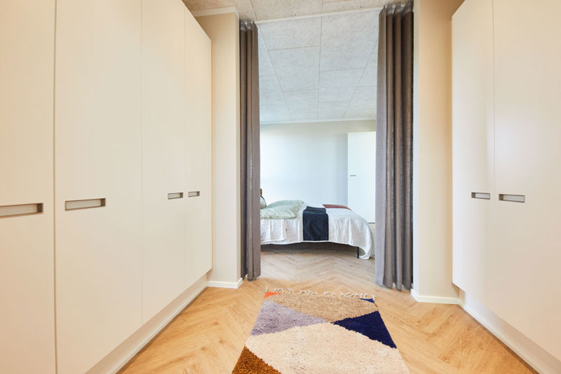 Walk-in passage fra eurodan-huse med lyse skabsvægge på begge sider, lange integrerede greb, gardiner som rumdeling, farvet gulvtæppe og kig ind til soveværelse med seng – inspiration til garderobe mellem rum i nybygget hus og moderne husbyggeri.