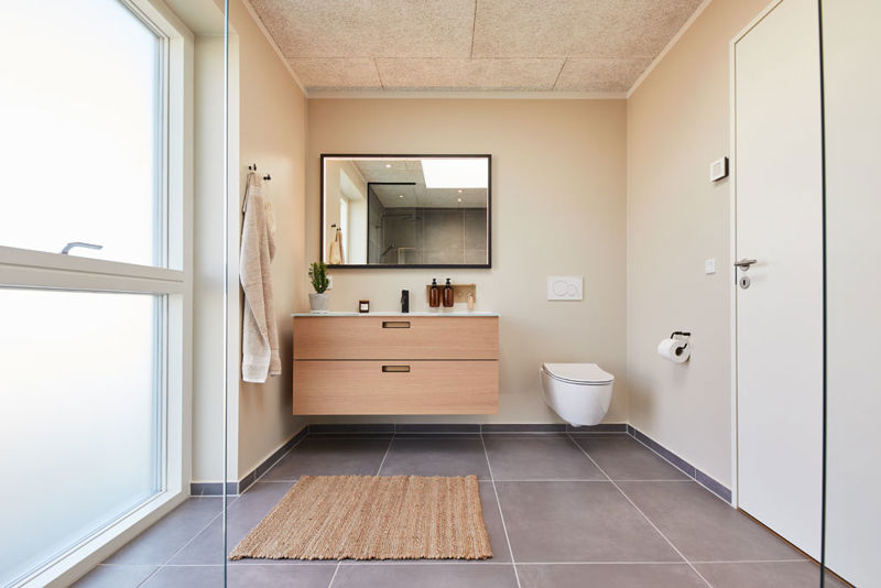 Moderne badeværelse i vinkelhus med svævende badmøbel i træ, stort spejl, væghængt toilet, mørke gulvfliser og stort vinduesparti – inspiration til nybyg og drømmehuset.