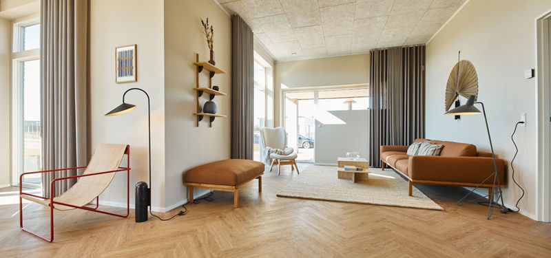 Stue fra eurodan-huse med cognacfarvet sofa, matchende bænk, rød loungestol, sort standerlampe, træhylder på væggen, beige gulvtæppe, høje gardiner og store vinduer – inspiration til nybyggeri, husbyggeri og moderne stueindretning.