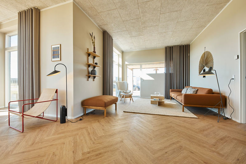 Stue fra eurodan-huse med cognacfarvet sofa, matchende bænk, rød loungestol, sort standerlampe, træhylder på væggen, beige gulvtæppe, høje gardiner og store vinduer – inspiration til nybyggeri, husbyggeri og moderne stueindretning.