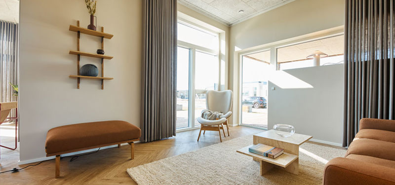 Stue fra eurodan-huse med cognacfarvet sofa, lys lænestol ved stort vindue, træbænk under væghylder, beige gulvtæppe, lavt sofabord og udgang mod terrasse – inspiration til nybyggeri, husbyggeri og stue med tæt kontakt til uderum.