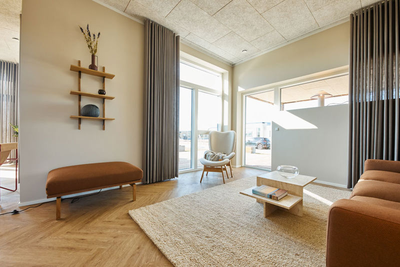 Stue fra eurodan-huse med cognacfarvet sofa, lys lænestol ved stort vindue, træbænk under væghylder, beige gulvtæppe, lavt sofabord og udgang mod terrasse – inspiration til nybyggeri, husbyggeri og stue med tæt kontakt til uderum.