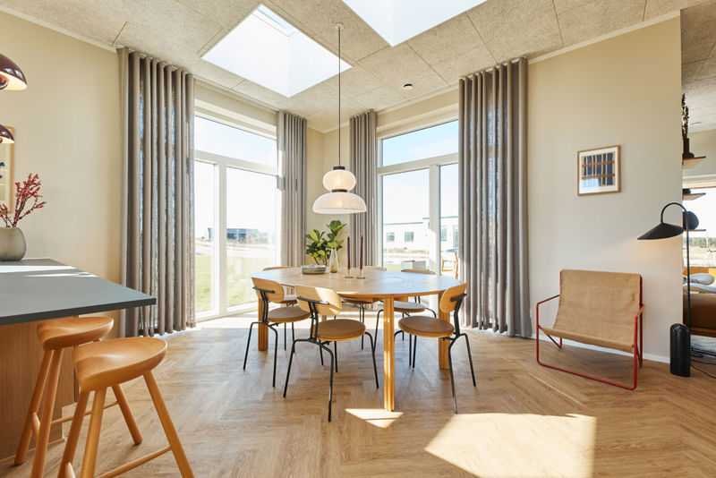 Lyst køkken-alrum i et moderne nybyggeri med rundt spisebord, store vinduespartier, ovenlys, lange gardiner og sildebensgulv.
