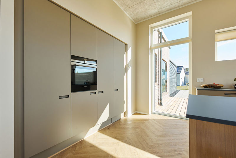 Moderne køkken i vinkelhus med høj skabsvæg, integreret ovn, sildebensgulv og åben terrassedør – inspiration til nybyg og drømmehuset.