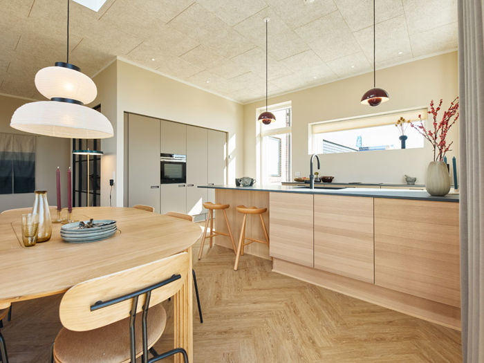 Lyst køkken-alrum i moderne vinkelhus med træfronter, køkkenø, barstole, spiseplads og dekorative pendellamper – inspiration til nybyg og drømmehuset.
