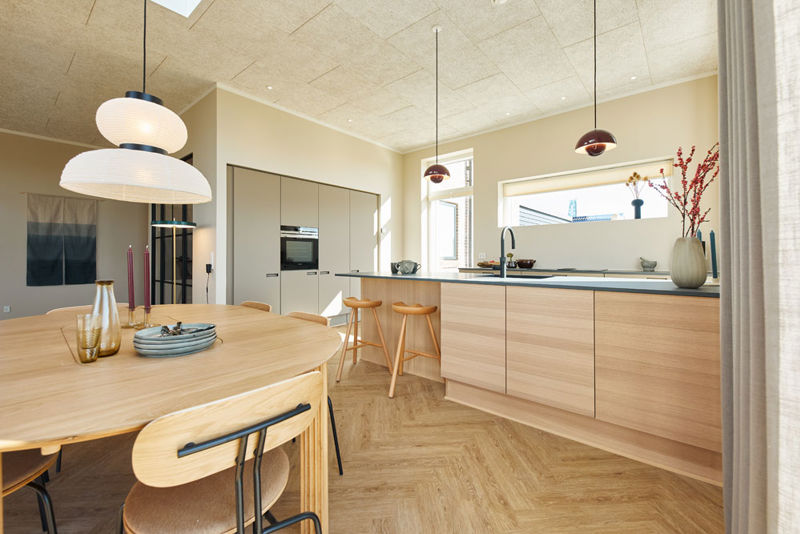 Lyst køkken-alrum i moderne vinkelhus med træfronter, køkkenø, barstole, spiseplads og dekorative pendellamper – inspiration til nybyg og drømmehuset.