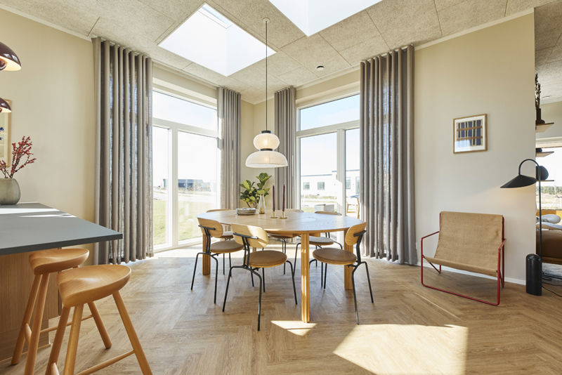 Lyst køkken-alrum fra eurodan-huse med rundt træspisebord, træstole, sildebensgulv, store vinduer med lange gardiner, ovenlys i loftet, pendel over spisepladsen, køkkenø med barstole og loungehjørne i siden – inspiration til nybyggeri med varme materialer og masser af dagslys.