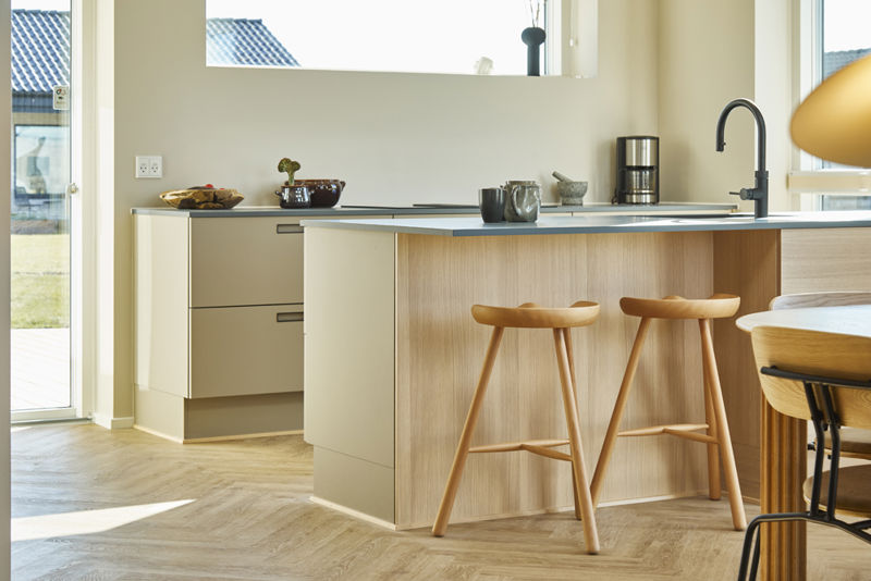 Detalje fra lyst køkken-alrum fra eurodan-huse med køkkenø i lyst træ, to træbarstole, sort armatur, vindueslys, hvide underskabe, kaffemaskine på bordpladen og rundt spisebord tæt ved køkkenet – inspiration til varme materialer, praktisk køkkenø og moderne nybyggeri.