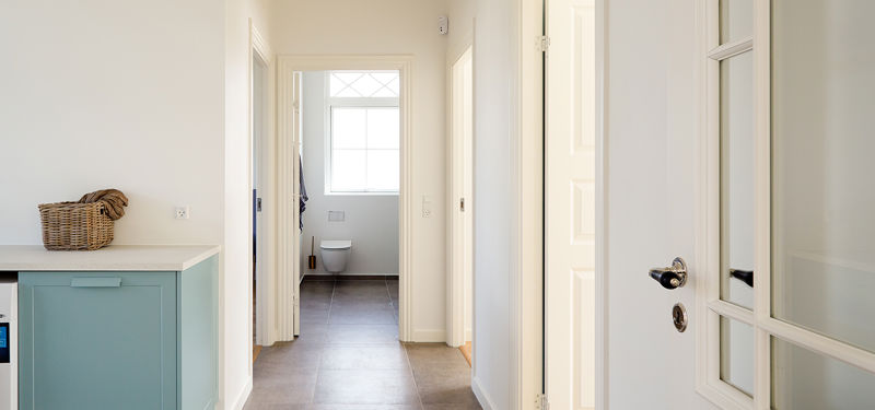Gang fra eurodan-huse med lyse vægge, fyldningsdøre, grå gulvfliser, kig til badeværelse, sprosset dør i forgrunden og turkis skab i siden – inspiration til klassisk nybyggeri, palæstil og sammenhængende boligindretning.