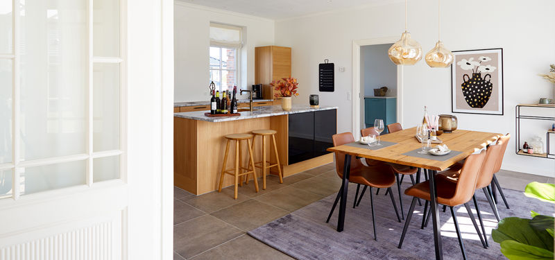 Spiseplads og køkken fra eurodan-huse med træbord, brune stole, gyldne glaspendler, køkkenø med træbarstole, mørke underskabe og klassisk hvid dør i forgrunden – inspiration til palæstil, nybyggeri og varme husdrømme.