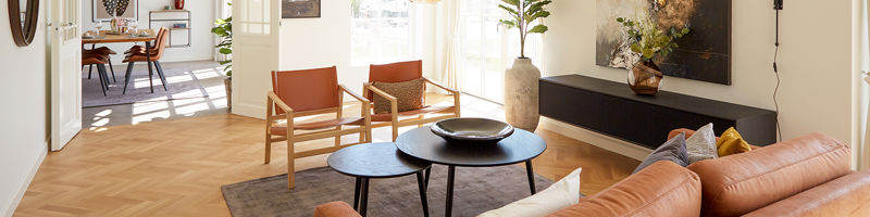 Lys stue fra eurodan-huse med cognacfarvet sofa, to læderlænestole, sorte runde sofaborde, stor plantekrukke, lav sort skænk, kunst på væggen og dobbeltdør mod spiseplads – inspiration til nybyggeri, husbyggeri og sammenhængende opholdsrum.
