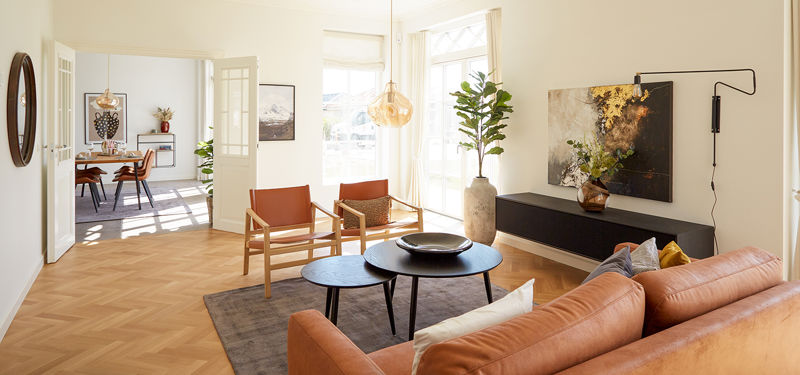 Lys stue fra eurodan-huse med cognacfarvet sofa, to læderlænestole, sorte runde sofaborde, stor plantekrukke, lav sort skænk, kunst på væggen og dobbeltdør mod spiseplads – inspiration til nybyggeri, husbyggeri og sammenhængende opholdsrum.