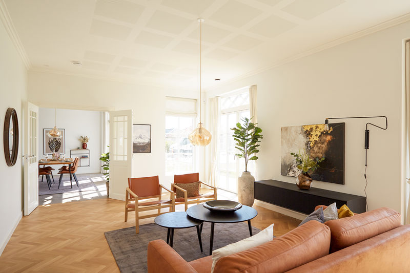 Lys stue fra eurodan-huse med cognacfarvet sofa, to læderlænestole, sorte runde sofaborde, stor plantekrukke, lav sort skænk, kunst på væggen og dobbeltdør mod spiseplads – inspiration til nybyggeri, husbyggeri og sammenhængende opholdsrum.