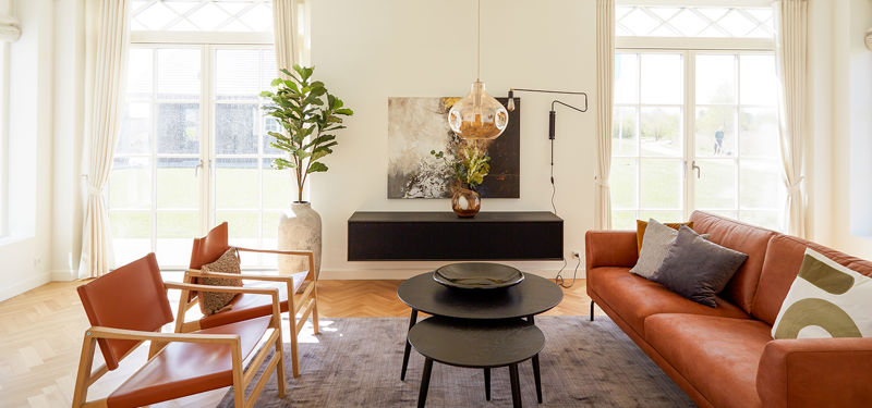 Stue fra eurodan-huse med cognacfarvet sofa, to lænestole i samme varme tone, sorte runde sofaborde, mørk lav skænk, stor plante i krukke og lyse vinduer med klassiske sprosser – inspiration til nybyggeri, husbyggeri og husdrømme med karakter.