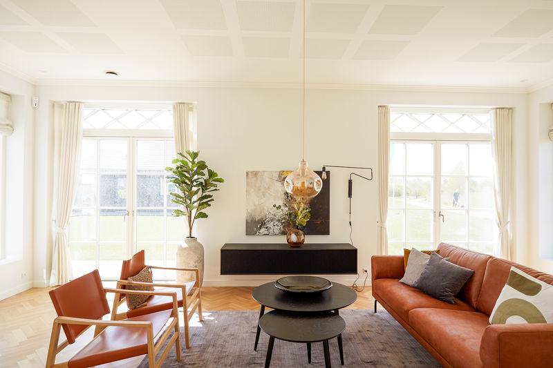 Stue fra eurodan-huse med cognacfarvet sofa, to lænestole i samme varme tone, sorte runde sofaborde, mørk lav skænk, stor plante i krukke og lyse vinduer med klassiske sprosser – inspiration til nybyggeri, husbyggeri og husdrømme med karakter.