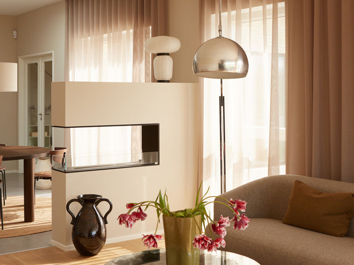 Hyggelig opholdsstue i moderne nybyg med sofa, rundt glasbord, blomster, gulvlampe og lange gardiner samt kig mod spisepladsen – inspiration til et lyst drømmehus.