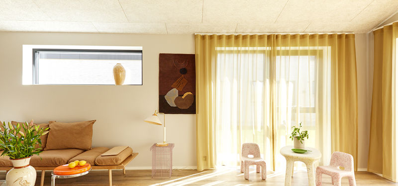 Farverig stue fra eurodan-huse med lav daybed, lille sidebord, grøn plante, børnestole, lyse gardiner, stort vinduesparti og blødt sollys på gulvet – inspiration til personligt nybyggeri, husbyggeri og kreative husdrømme.