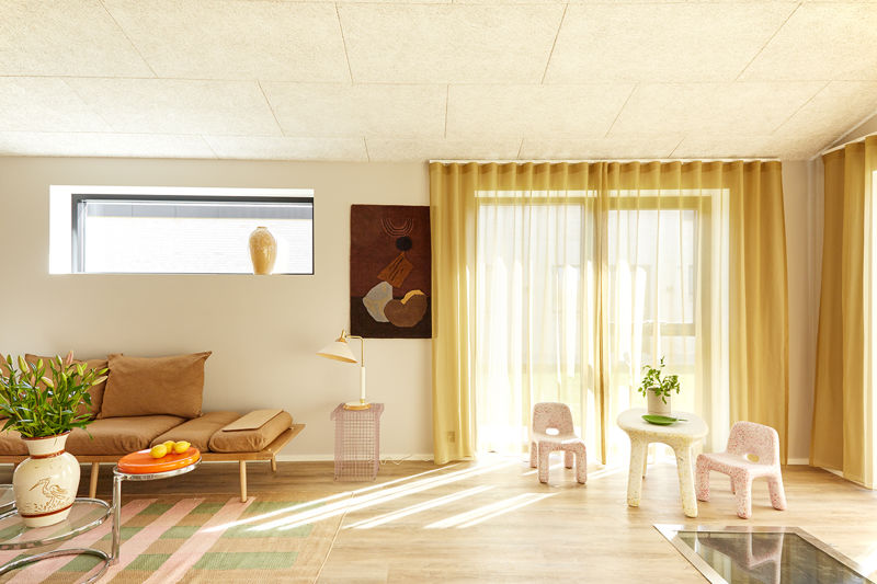 Farverig stue fra eurodan-huse med lav daybed, lille sidebord, grøn plante, børnestole, lyse gardiner, stort vinduesparti og blødt sollys på gulvet – inspiration til personligt nybyggeri, husbyggeri og kreative husdrømme.