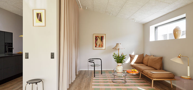 Stue fra eurodan-huse med brun daybed, lavt bord med appelsiner, mønstret tæppe, gul læselampe, lange rosa gardiner, lille sort stol og kig til sort køkken – inspiration til nybyggeri, husbyggeri og personlig indretning af stue.