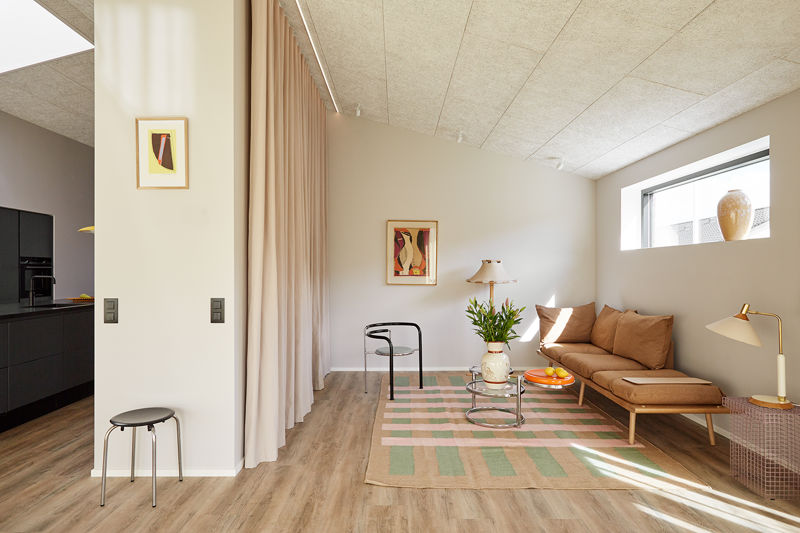 Stue fra eurodan-huse med brun daybed, lavt bord med appelsiner, mønstret tæppe, gul læselampe, lange rosa gardiner, lille sort stol og kig til sort køkken – inspiration til nybyggeri, husbyggeri og personlig indretning af stue.