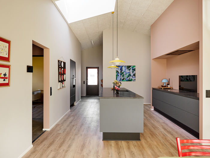 Køkken-alrum fra eurodan-huse med lang køkkenø i midten, mørk bordplade, rosa væg og indrammet køkkenniche, mørke underskabe, sort skabsvæg, ovenlys i kiploft, farverig kunst og rød stol i forgrunden – inspiration til personligt nybyggeri og unikke husdrømme.