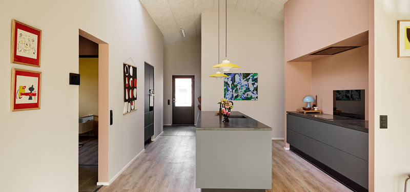 Køkken-alrum fra eurodan-huse med lang køkkenø i midten, mørk bordplade, rosa væg og indrammet køkkenniche, mørke underskabe, sort skabsvæg, ovenlys i kiploft, farverig kunst og rød stol i forgrunden – inspiration til personligt nybyggeri og unikke husdrømme.