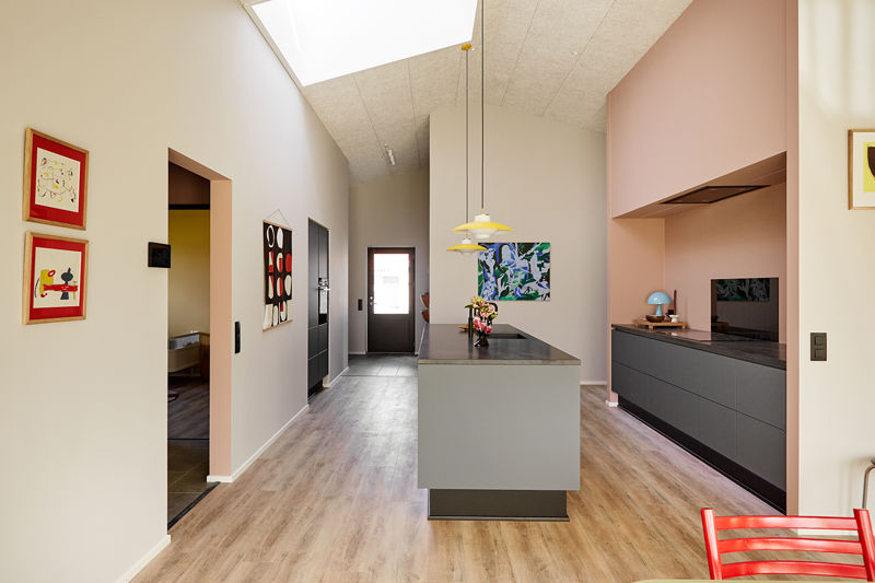 Køkken-alrum fra eurodan-huse med lang køkkenø i midten, mørk bordplade, rosa væg og indrammet køkkenniche, mørke underskabe, sort skabsvæg, ovenlys i kiploft, farverig kunst og rød stol i forgrunden – inspiration til personligt nybyggeri og unikke husdrømme.