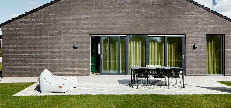 Bred terrasse foran murstensfacade fra eurodan-huse med store skydedøre, sorte udendørslamper, spisebord med mørke stole, loungepuf på flisebelægning og grøn plæne i forgrunden – inspiration til funktionelt nybyggeri og udeliv tæt på hjemmet.