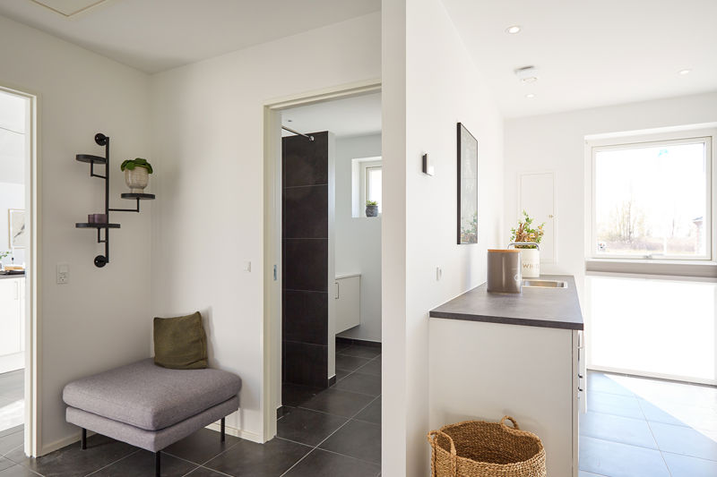 Entréområde fra eurodan-huse med lille bænk, væghylde i sort metal, plante i krukke, mørke gulvfliser, åbning til badeværelse, bordplade med vase og stor vinduesdør mod haven – inspiration til praktisk opbevaring, entréindretning, nybyggeri og moderne husbyggeri.