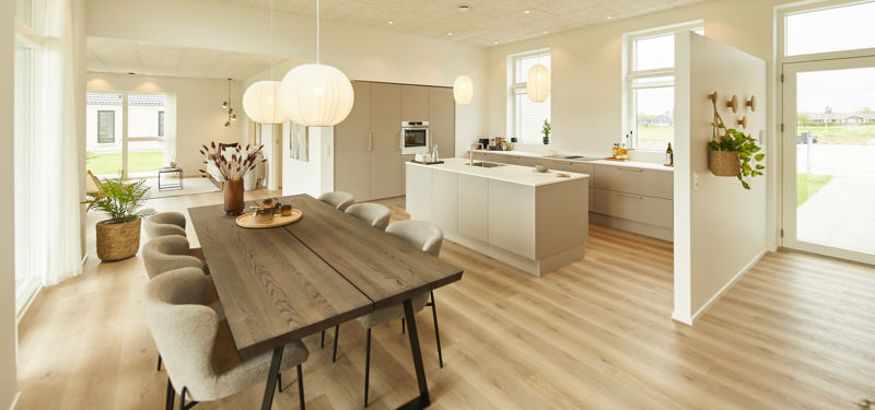 Åbent køkken-alrum fra eurodan-huse med langt spisebord, lyse polstrede stole, runde pendler, hvid køkkenø, lyse skabsfronter, vinduer på flere sider, ovenlys i loftet og direkte kig til opholdsrum – inspiration til nybyggeri med stor rumfornemmelse.