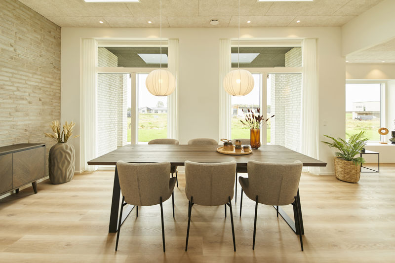 Spiseplads i køkken-alrum fra eurodan-huse med langt mørkt spisebord, seks polstrede stole, to runde pendler, store vinduespartier med lyse gardiner, lys murstensvæg, sideboard med vase og grøn plante i hjørnet – inspiration til nybyggeri, husbyggeri og moderne husdrømme.
