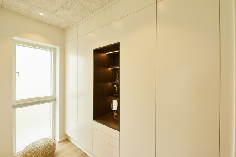 Indbygget garderobevæg fra eurodan-huse med hvide fronter, stort mørkt nichefelt med åbne hylder, skuffer under nichen, integreret hyldelys, lyst trægulv og vindue ved siden af – inspiration til specialdesignet walk-in closet i nybyggeri og husdrømme.