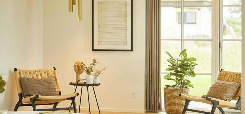Hyggeligt stuehjørne fra eurodan-huse med to flettede loungestole, sort rundt sidebord, grøn plante i kurv, vægkunst, messingfarvede pendler og stort vindue med lyse gardiner – inspiration til nybyggeri, husbyggeri og rolige husdrømme.