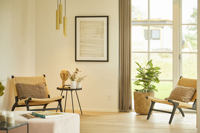 Hyggeligt stuehjørne fra eurodan-huse med to flettede loungestole, sort rundt sidebord, grøn plante i kurv, vægkunst, messingfarvede pendler og stort vindue med lyse gardiner – inspiration til nybyggeri, husbyggeri og rolige husdrømme.