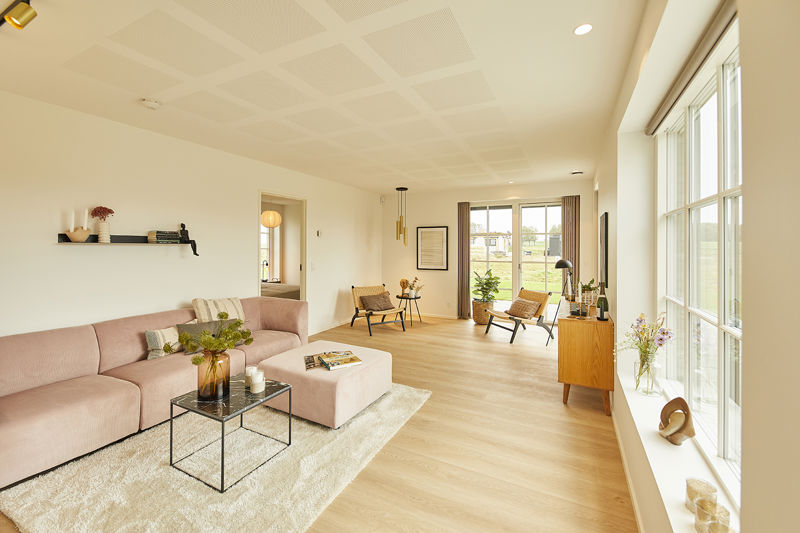 Lang stue fra eurodan-huse med rosa sofa og puf, sort glasbord, lyst tæppe, stort vinduesparti, træskænk, flettede loungestole i baggrunden og akustikloft – inspiration til nybyggeri, husbyggeri og lyse opholdsrum.