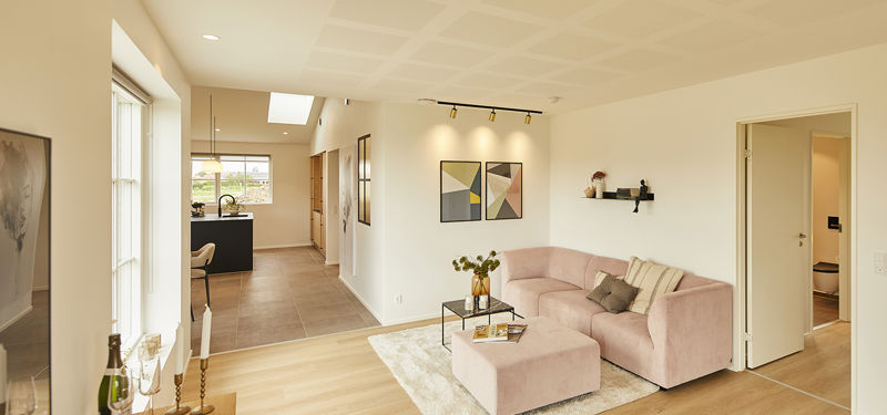 Stue fra eurodan-huse med rosa modulsofa og puf, sort sofabord, lyst tæppe, spotbelysning over vægkunst, åben forbindelse til køkken og dør mod værelse – inspiration til nybyggeri, husbyggeri og funktionelle opholdsrum.