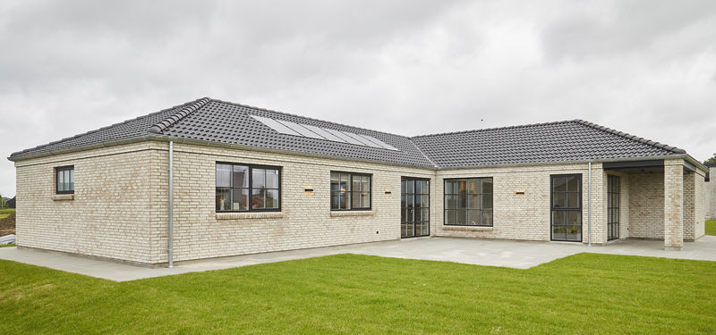 Udvendig facade fra eurodan-huse med lyse mursten, mørkt tegltag, sorte sprossede vinduer, overdækket indgangsparti, stor flisebelagt terrasse og grøn græsplæne – inspiration til palæstil, nybyggeri og klassisk husbyggeri.