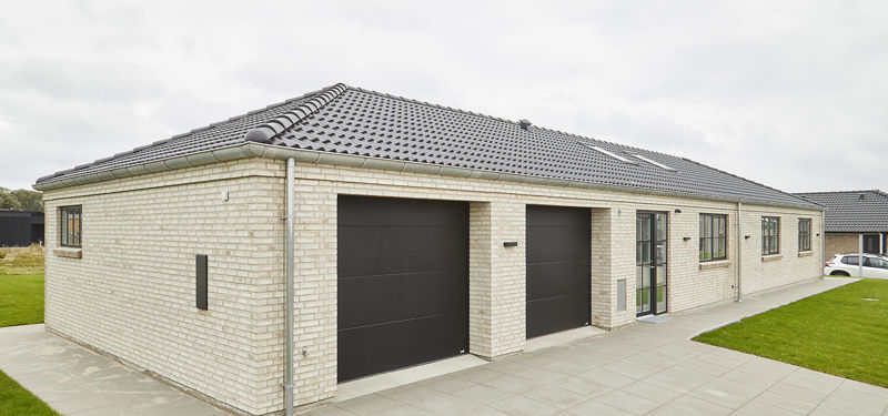 Facade fra eurodan-huse med lyse mursten, sort tegltag, to mørke garageporte, sprossede vinduer, terrassedør og bred flisegang langs huset – inspiration til klassisk palæstil, praktisk nybyggeri og husdrømme med garage integreret i arkitekturen.