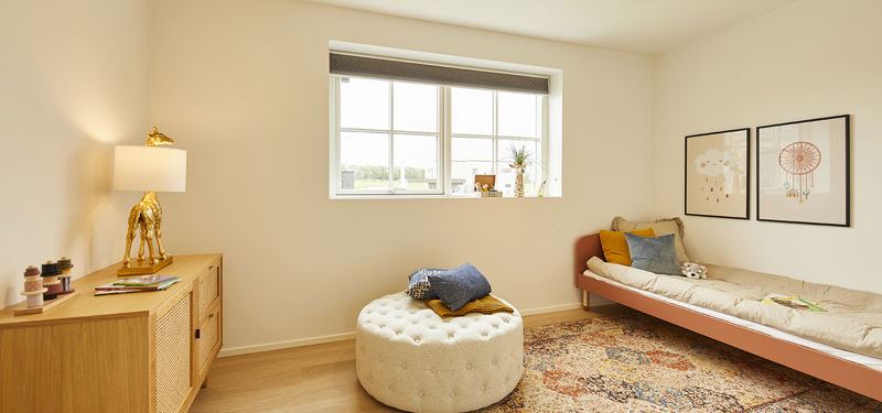 Lyst børneværelse fra eurodan-huse med bredt sprosset vindue, daybed med puder, rund polstret puf, farverigt tæppe, lav trækommode, lampe med giraffod og plakater på væggen – inspiration til børneværelser i nybyggeri, husbyggeri og moderne husdrømme.